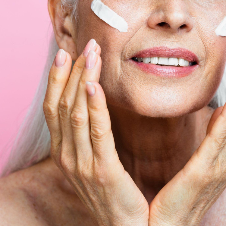 Mulher a sorrir enquanto aplica creme facial nas maçãs do rosto sobre um fundo rosa.